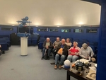 Foto sedících členů se záběrem na přístroj, který promítá oblohu