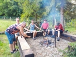 Pět členů SONS sedí na lavičkách okolo ohně a pečou masové špízy