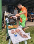 Na obrázku vrchní kuchař Aleš, má na sobě slušivou zástěru s nápisem „Tady vařím já“ 😊. Stojí před grilem a otáčí klobásy. Na stole leží plno dalších klobás připravených do akce 😊. 