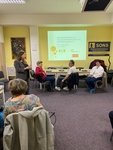 V místnosti kolem stolů sedí lidé, žena jim přednáší a na zeď je promítána prezentace o manipulaci a podvodném jednání.