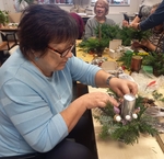 Na obrázku jedna z účastnic dílniček při práci na svém svícnu – základ ze zelených větviček, uprostřed stříbrná svíčka a okolo ní paní upevňuje stříbrné, bílé a fialové dekorace. Na obrázku jedna z účastnic dílniček při práci na svém svícnu – základ ze zelených větviček, uprostřed stříbrná svíčka a okolo ní paní upevňuje stříbrné, bílé a fialové dekorace.