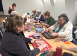 Celkový pohled na naši skupinu - sedíme u dlouhého stolu, na kterém je pracovní nepořádek a tvoříme výrobky z plstě. 