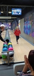 Popis fotografie: Na fotografii je zachycená jednoduchá, veselá scéna z bowlingu. V popředí je vidět stojan s barevnými bowlingovými koulemi. Jedna žena v červeném tričku právě odchází od dráhy. Další člověk stojí u koulí a vybírá si tu svoji.