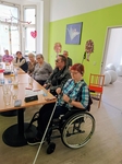Na fotografii je skupina lidí sedících okolo stolu. V popředí sedí žena na invalidním vozíku. V ruce drží bílou hůl WeWalk