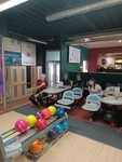 Na fotografii je velký bowlingový sál. V popředí jsou stojany s barevnými bowlingovymi koulemi. V pozadí jsou stoly a židle u kterých sedí lidé a sledují hru